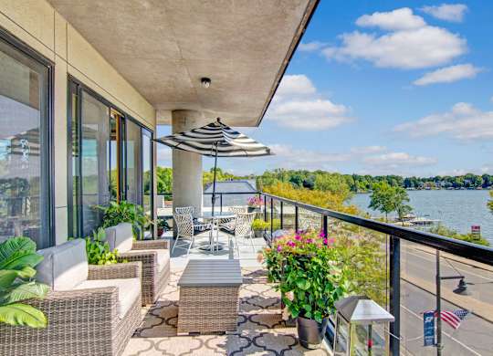 A private balcony here at The Mist featuring comfortable outdoor seating with woven chairs, a small table, and potted greenery arranged along a patterned outdoor rug. A glass railing frames expansive lake and treetop views, while a striped umbrella and sliding glass doors create an inviting space to relax outdoors and enjoy fresh air and natural scenery.