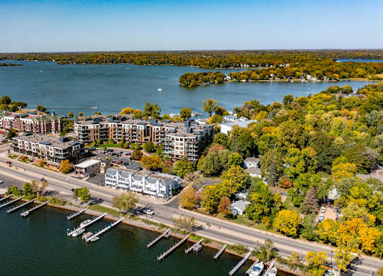 A sweeping aerial view here at The Mist showcasing a waterfront apartment community set along a scenic lake, with modern buildings lining the shoreline, nearby docks extending into the water, and tree-filled neighborhoods surrounding the area. Expansive blue water, winding roads, and lush greenery create a vibrant lakeside setting with picturesque views in every direction.