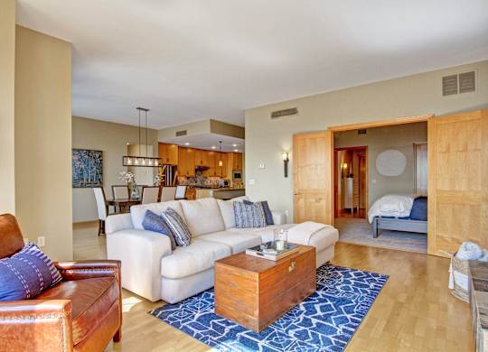 A spacious open living room here at The Mist featuring light wood-style flooring, a plush sectional sofa with accent pillows, and a wood coffee table set on a patterned area rug. The space flows into a dining area and kitchen with warm wood cabinetry and pendant lighting, while sliding doors open to a bedroom, creating a bright, inviting layout ideal for everyday living and entertaining.