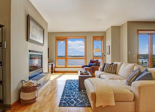 A bright living room here at The Mist featuring light wood-style flooring, a comfortable sectional sofa with textured accent pillows, and a patterned area rug centered around a sleek coffee table. A built-in fireplace adds warmth along one wall, while large windows frame scenic water views and fill the space with natural light.