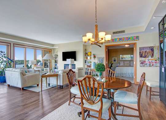 A spacious open living and dining area here at The Mist featuring wood-style flooring, a round dining table with classic wood chairs, and a comfortable seating area with armchairs and side tables. Large windows line the space, bringing in abundant natural light and scenic water views, while built-in shelving, artwork, and warm lighting create an inviting atmosphere.