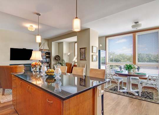 A spacious open kitchen and dining area here at The Mist featuring a large island with bar seating, smooth countertops, and warm wood cabinetry beneath modern pendant lighting. The layout opens to a dining nook with a round table near wide windows, allowing natural light to brighten the space and create an inviting setting for meals and gatherings.
