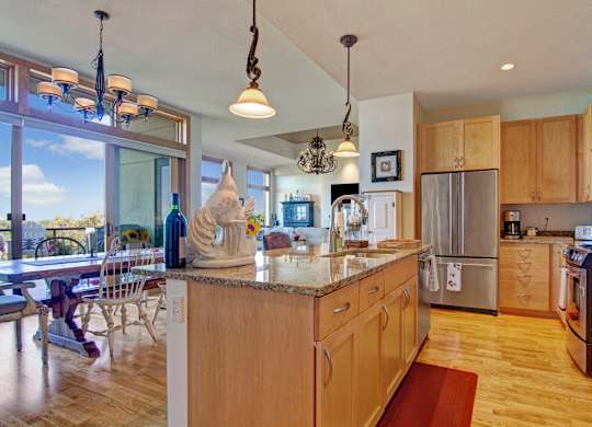 A bright open kitchen here at The Mist featuring a large central island with smooth countertops, warm wood cabinetry, and a modern sink with a tall faucet. Pendant and chandelier lighting add character overhead, while the layout flows into a dining area with large windows, hardwood-style flooring, and views that bring in natural light.