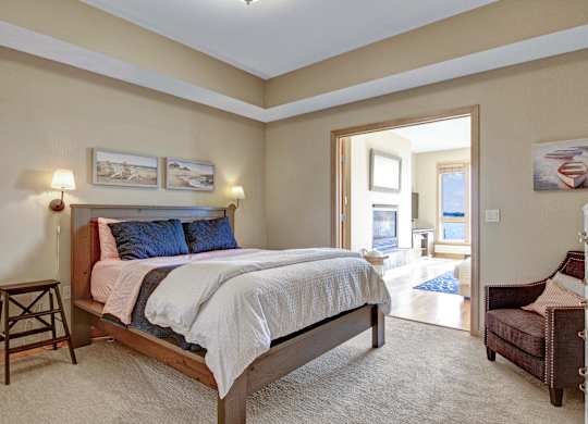 A serene bedroom here at The Mist featuring a spacious layout with a wood-framed bed dressed in soft neutral bedding, matching wall-mounted lamps, and framed coastal artwork above the headboard. Plush carpeting adds comfort underfoot, while an open doorway leads to a bright adjoining living area with large windows that bring in abundant natural light and scenic views.