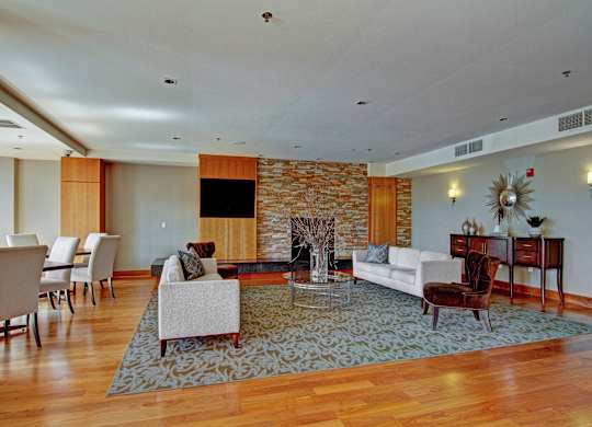 A welcoming community lounge here at The Mist featuring an open layout with plush seating arranged around a glass coffee table, warm wood-style flooring, and a textured accent wall with a fireplace. A long dining table with upholstered chairs sits nearby, while soft lighting and neutral décor create an inviting space for relaxing or gathering.
