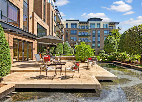 A scenic outdoor patio here at The Mist featuring café-style tables and chairs arranged on a spacious stone terrace overlooking a tranquil water feature. Lush landscaping, manicured trees, and surrounding apartment buildings frame the setting, creating a peaceful space for relaxing outdoors while enjoying fresh air and sunny views.
