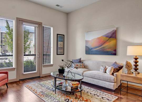 A stylish living room here at Residence at the COR featuring wood-style flooring, a light neutral sofa with patterned accent pillows, a glass coffee table on a colorful area rug, and modern wall art, with large windows and a glass door bringing in natural light and creating a bright, inviting space for relaxing or entertaining.