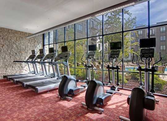 A bright fitness center here at Residence at the COR showcasing a lineup of treadmills and elliptical machines arranged along expansive floor-to-ceiling windows, allowing natural light and views of the landscaped courtyard and pool, with warm carpeted flooring, textured stone accent walls, and an open, motivating layout designed for comfortable daily workouts.