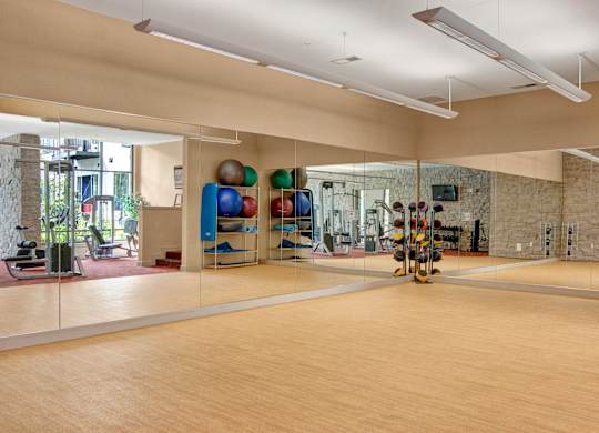A spacious fitness studio here at Residence at the COR featuring a wide open workout area with smooth flooring, full mirrored walls, and overhead lighting, complemented by neatly organized exercise balls, mats, and free weights, with adjacent cardio and strength equipment visible through the mirrors, creating a versatile space ideal for stretching, group classes, and functional training.