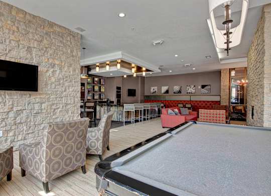 A stylish resident lounge here at Residence at the COR featuring a spacious open layout with a pool table, patterned lounge chairs, and a mounted television, complemented by stone accent walls, warm wood finishes, a modern bar with seating, cozy sofas, and contemporary lighting that creates an inviting space for relaxing, socializing, and entertainment.