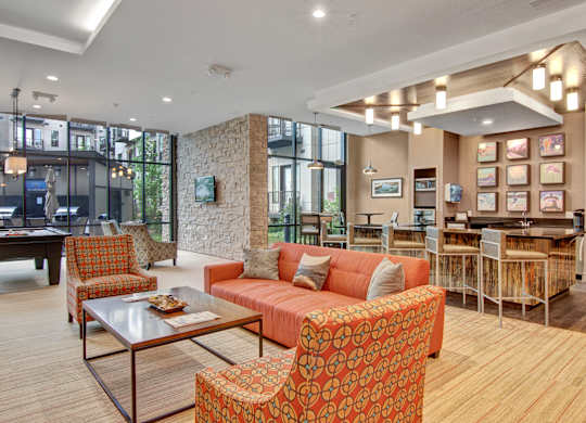 A contemporary resident lounge here at Residence at the COR showcasing a bar-height island with cushioned stools and sleek countertops, complemented by warm wood accents, an orange tufted banquette, modern wall art, and layered recessed and pendant lighting, all within an open, inviting layout designed for socializing, entertaining, and everyday relaxation.