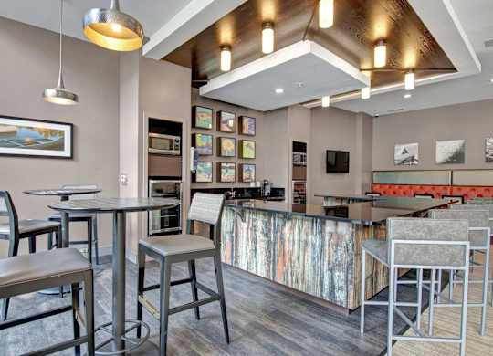A stylish resident lounge here at Residence at the COR featuring a bar-height island with sleek countertops and modern stools, complemented by café-style tables, warm wood ceiling accents, contemporary pendant lighting, colorful framed artwork, and an inviting seating area that creates a polished, social setting for gathering, entertaining, or unwinding.