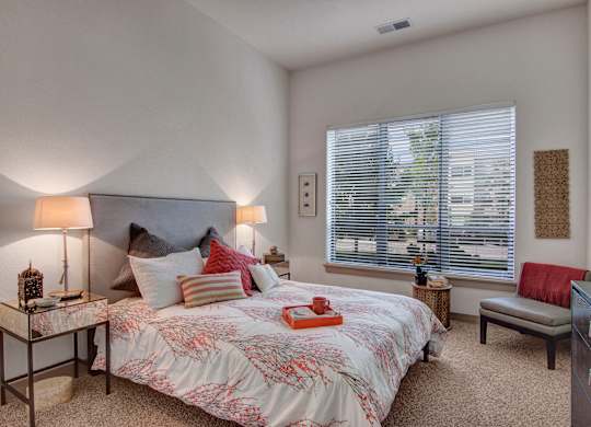 A serene bedroom here at Residence at the COR featuring a plush bed with layered neutral and accent pillows, a patterned comforter, and matching bedside tables with soft-glow lamps, complemented by cozy carpet, a large window with blinds letting in natural light, and thoughtfully placed seating and decor that create a calm, inviting retreat.