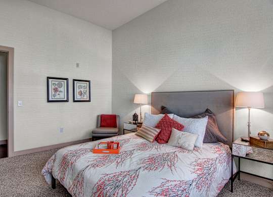 A cozy bedroom here at Residence at the COR showcasing a comfortable bed with layered neutral and warm accent pillows, a patterned comforter, and matching bedside tables with soft lighting, complemented by plush carpeting, simple wall art, and a small seating area that creates a calm, welcoming space designed for everyday comfort and relaxation.