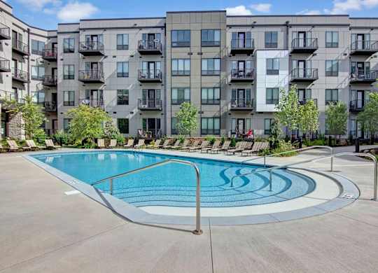 A resort-style outdoor pool here at Residence at the COR featuring a curved entry with handrails and shallow steps, surrounded by a wide concrete deck lined with lounge chairs, while a modern apartment building with private balconies and landscaped greenery creates a polished, inviting backdrop for relaxation and social time.