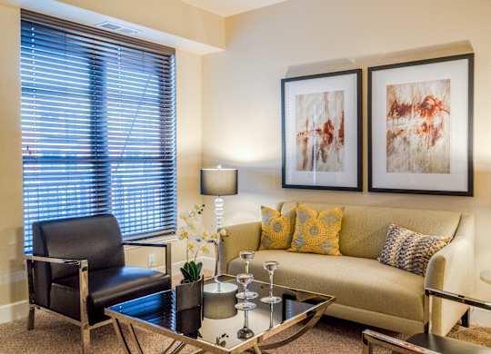 A warm and inviting living room here at Siena featuring a comfortable sofa accented with patterned throw pillows, a sleek glass coffee table, and modern table lamps that provide soft lighting, complemented by contemporary wall art, a cozy accent chair, and a large window with blinds that brings in natural light and creates a relaxed, welcoming atmosphere.