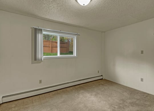 Empty bedroom here at Montclair Apartments with beige carpet and off-white walls, featuring a single window with vertical blinds and a view of a wooden fence and greenery outside. Neutral and calm atmosphere.