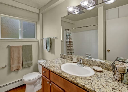 A modern bathroom here at Montclair Apartments with a granite countertop and sink, adjacent to a toilet. Towels hang by a frosted window, and soft, warm lighting creates a cozy ambiance.