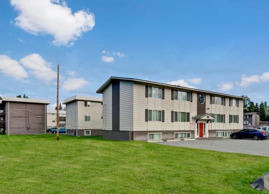 Two-story Montclair apartment building with white siding and red door set against a blue sky with fluffy clouds. Surrounding green lawn adds a serene touch.