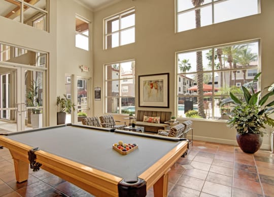 Bright recreation room here at Mozaic at Steele Park Apartments with a pool table, modern seating, large potted plants, and expansive windows showcasing a view of palm trees and a pool.