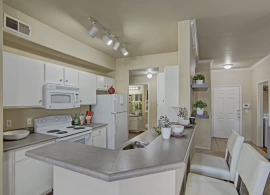 Modern kitchen here at Mozaic at Steele Park Apartments with white cabinets, gray countertops, and bar seating. Bright lighting, decorative plants, and a cozy, welcoming ambiance.