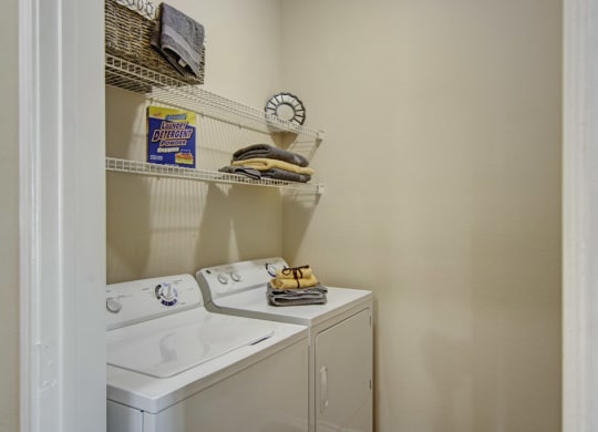 Compact laundry room here at Mozaic at Steele Park Apartments with a washing machine and dryer. Shelves hold detergent, a wicker basket, and neatly folded towels, creating an organized feel.