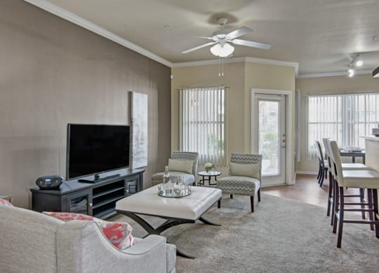 Modern living room here at Mozaic at Steele Park Apartments with neutral tones, featuring a TV on a stand, a gray sofa, patterned chairs, bar stools, and tall windows letting in natural light.