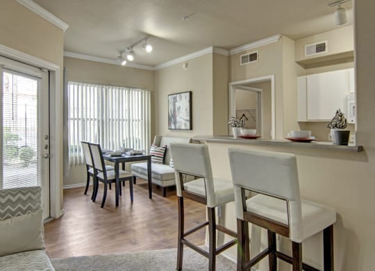 A cozy apartment interior here at Mozaic at Steele Park Apartments with a dining area and kitchen. The dining area has a black table and chairs by large windows. The kitchen features a breakfast bar with high chairs, evoking a warm, inviting atmosphere.