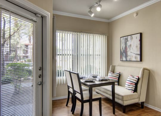 Cozy dining area here at Mozaic at Steele Park Apartments with a beige bench and chairs at a wooden table set for four. Large windows with vertical blinds let in natural light. Abstract art on the wall.