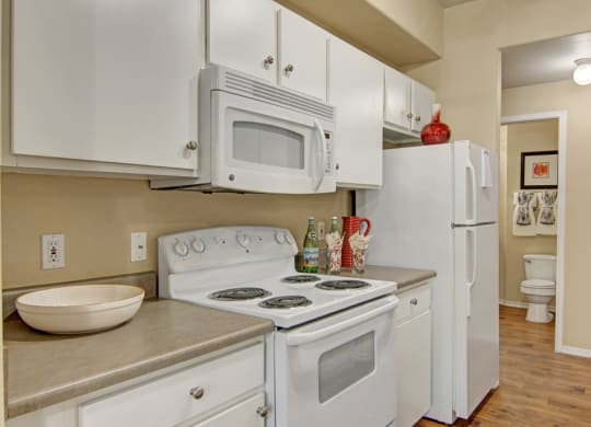 A cozy kitchen here at Mozaic at Steele Park with white cabinets, stove, and microwave. A red vase sits atop the fridge. A tidy bathroom with framed art is visible in the background.