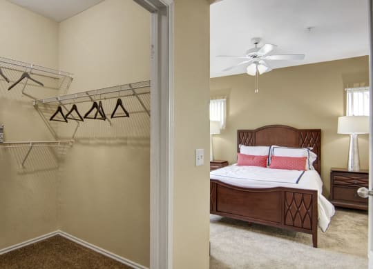 Bedroom view here at Mozaic at Steele Park Apartments from a closet entrance, featuring empty hangers on wire shelves. Warm tones, neatly made bed with red pillows, and serene ambiance.