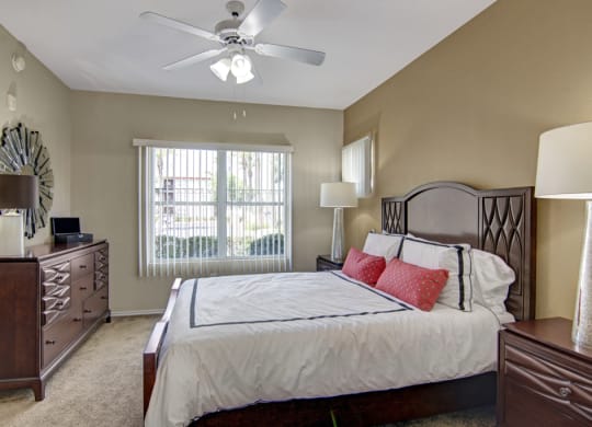 Cozy bedroom here at Mozaic at Steele Park Apartments with a large bed, white linens, and red accent pillows. A ceiling fan hangs above. Wooden furniture and large window create a warm atmosphere.