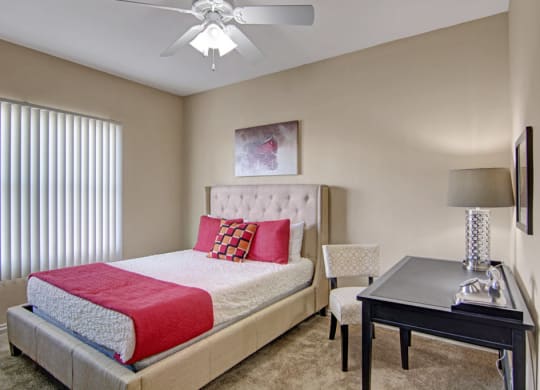 A cozy bedroom here at Mozaic at Steele Park Apartments with a beige tufted bed, white and red bedding, and a checkered chair. A black desk with a lamp and modern decor adds elegance.