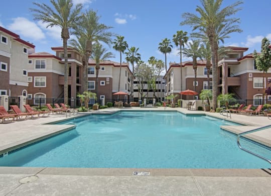 Our luxurious Mozaic at Steele Park Apartment complex pool area with crystal-clear blue water, surrounded by palm trees and red lounge chairs. Calm, inviting atmosphere under a sunny sky.
