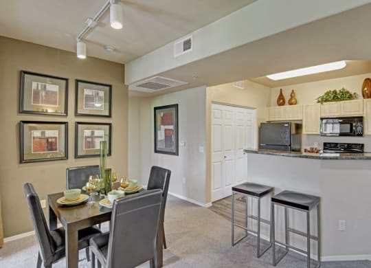 Modern dining and kitchen area here at Natura Villas Apartments with neutral tones. A dining table set for four, with green decor and artwork on the walls. Kitchen has bar stools, dark appliances, and decorative items on cabinets. Bright and inviting atmosphere.