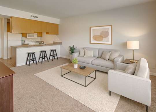 A bright, modern living room here at Confluence at Harvest Hills featuring an open-concept layout with a neutral sofa and armchair, a wooden coffee table on a light area rug, soft beige carpeting, a kitchen breakfast bar with stools, light wood cabinetry, a floor lamp, simple abstract wall art, and large windows allowing natural light to fill the space.