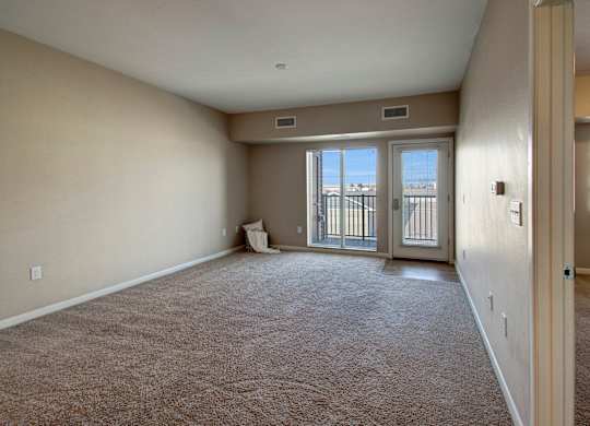 A spacious, unfurnished living room here at Dakota Commons featuring soft neutral walls, plush carpet flooring, and a sliding glass door with an adjacent exterior door that opens to a private balcony, allowing natural light to fill the open layout and highlight the room’s generous size.