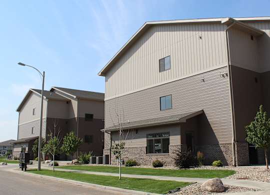A contemporary apartment building exterior here at Dakota Commons showcasing a multi-story structure with neutral two-tone siding, brick accents along the base, landscaped greenery, sidewalks, and a quiet residential street under a clear blue sky.
