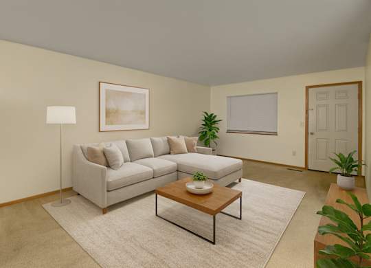 A bright, cozy living room here at Dakota featuring a light gray sectional sofa with neutral throw pillows, a wooden coffee table on a soft area rug, warm beige walls, indoor plants, simple wall art, and a floor lamp, creating a clean and inviting apartment living space.