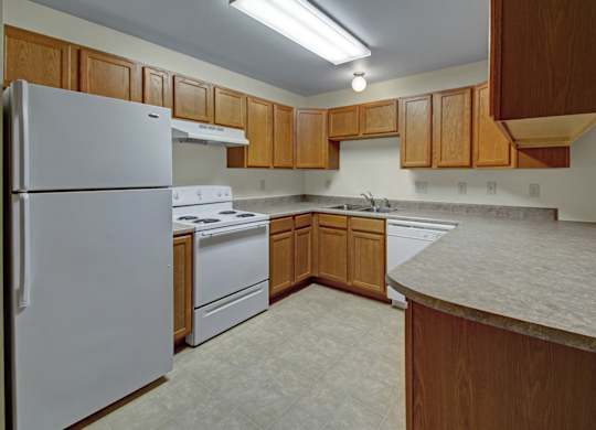 A bright, functional kitchen here at Dakota featuring warm wood cabinetry, white appliances including a refrigerator, stove, and dishwasher, durable neutral countertops, vinyl flooring, and ample counter space that creates a clean and practical apartment cooking area.