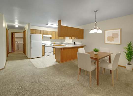 A spacious open-concept dining and kitchen area here at Dakota featuring warm wood cabinetry, a peninsula breakfast bar, white appliances, soft carpet transitioning to vinyl flooring, a four-seat dining table with modern chairs, and neutral lighting that creates a bright, welcoming apartment interior.