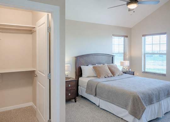 A bright, comfortable bedroom here at Dakota Ridge featuring a neatly made bed with neutral bedding, matching nightstands with lamps, soft carpeted flooring, a ceiling fan with light, two sunlit windows with blinds, and an open walk-in closet with built-in shelving for convenient storage.