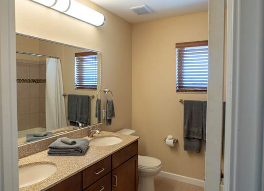 A clean, well-lit bathroom here at Dakota Ridge featuring a dual-sink vanity with granite countertops, dark wood cabinetry, a large wall mirror, neatly folded towels, a toilet, neutral wall tones, wood-style flooring, and a small window with blinds that brings in natural light.