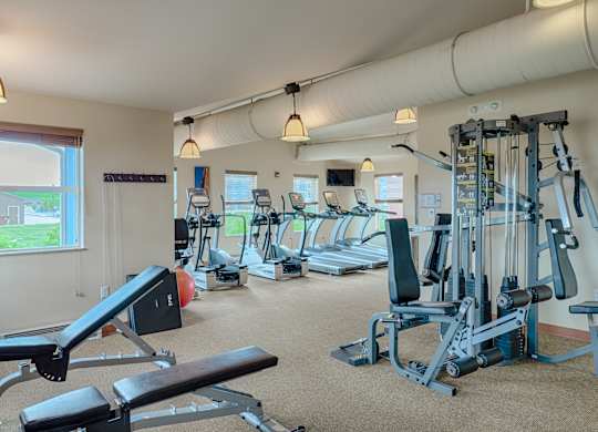A spacious, well-lit fitness center here at Dakota Ridge featuring treadmills lined along large windows, a multifunction weight machine, a padded workout bench, neutral carpet flooring, exposed ceiling ductwork, and warm pendant lighting that creates a clean, motivating environment for residents to exercise comfortably.