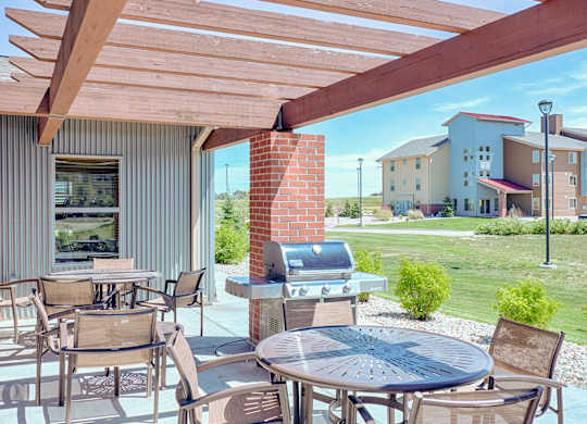 A shaded outdoor patio here at Dakota Ridge featuring a wooden pergola with exposed beams, round metal tables with mesh chairs, a stainless steel grill beside a brick column, and open views of manicured green lawns and nearby apartment buildings under a bright blue sky.