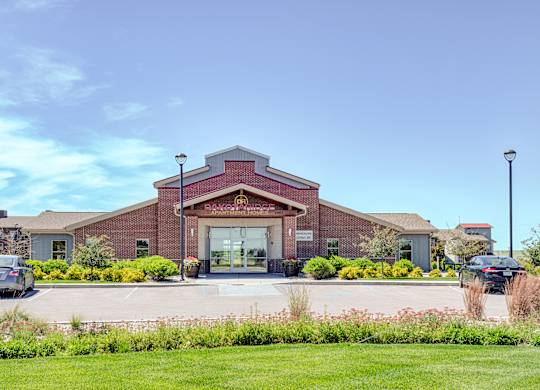 A welcoming exterior view here at Dakota Ridge featuring the brick clubhouse entrance with a peaked roofline, glass double doors, landscaped greenery, colorful planters, and a wide parking area set against a bright blue sky and well-maintained lawns.
