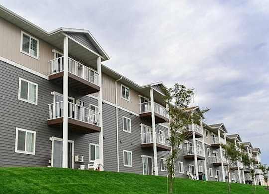 A modern apartment building exterior here at Pheasant Ridge showcasing multi-story residences with gray and beige siding, private balconies, white railings, evenly spaced windows, and a well-maintained green lawn with young trees under a lightly overcast sky.