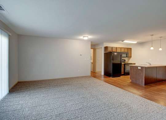 A spacious living room with a grey carpet and a kitchen area in the background.