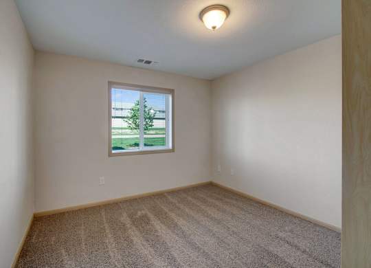 A bright bedroom here at Pheasant Ridge featuring plush neutral carpeting, soft beige walls with light wood trim, a centered window that brings in natural daylight and outdoor views, clean lines with ample wall space for furniture placement, and a simple ceiling light that creates a calm, comfortable atmosphere.