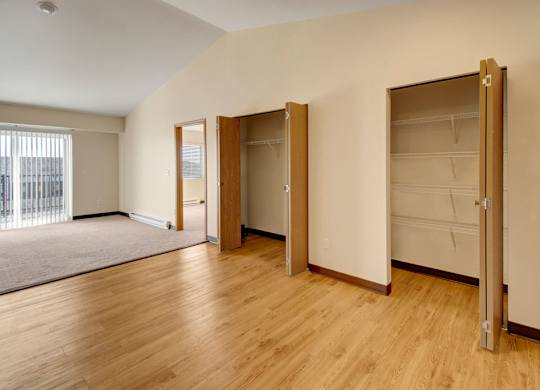 A spacious open living area here at Pheasant Ridge featuring warm wood-style flooring, multiple built-in closets with open shelving for storage, soft neutral walls, and a bright layout that flows into a carpeted living space with sliding glass doors that bring in natural light.